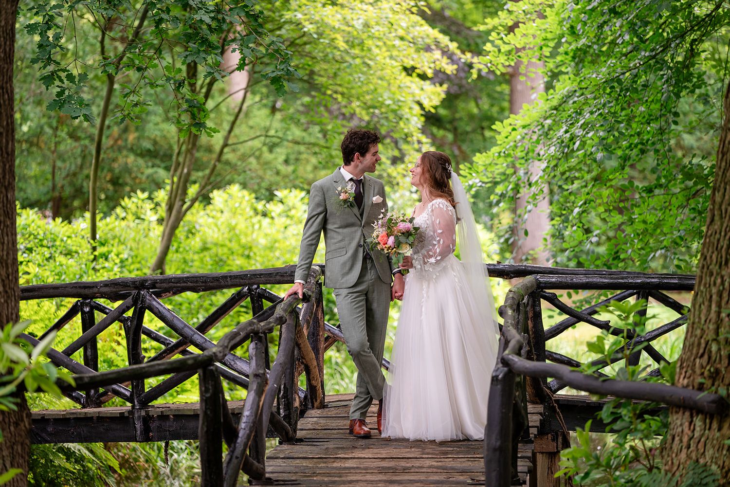 Bride and groom share a romantic moment on a rustic wooden bridge surrounded by lush green trees.