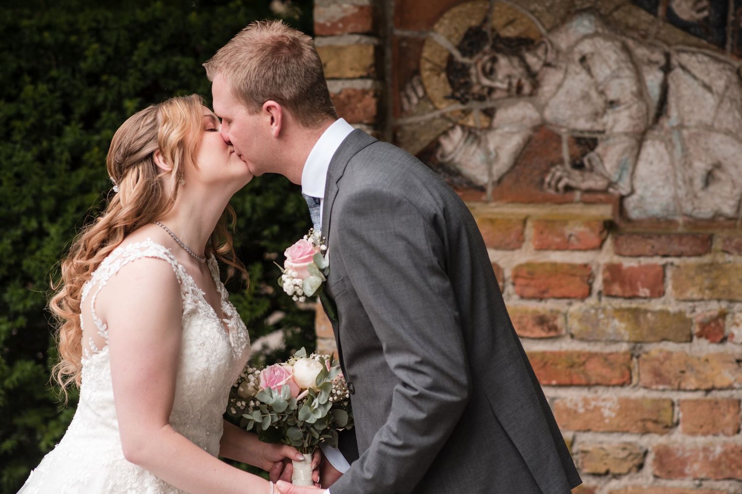 A bride in a lace wedding dress and a groom in a gray suit share a kiss beside a brick wall, holding a pink floral bouquet.