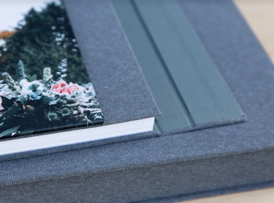 Close-up of a floral wedding photo album resting on a gray linen-covered presentation box with matching green fabric ribbon pulls.