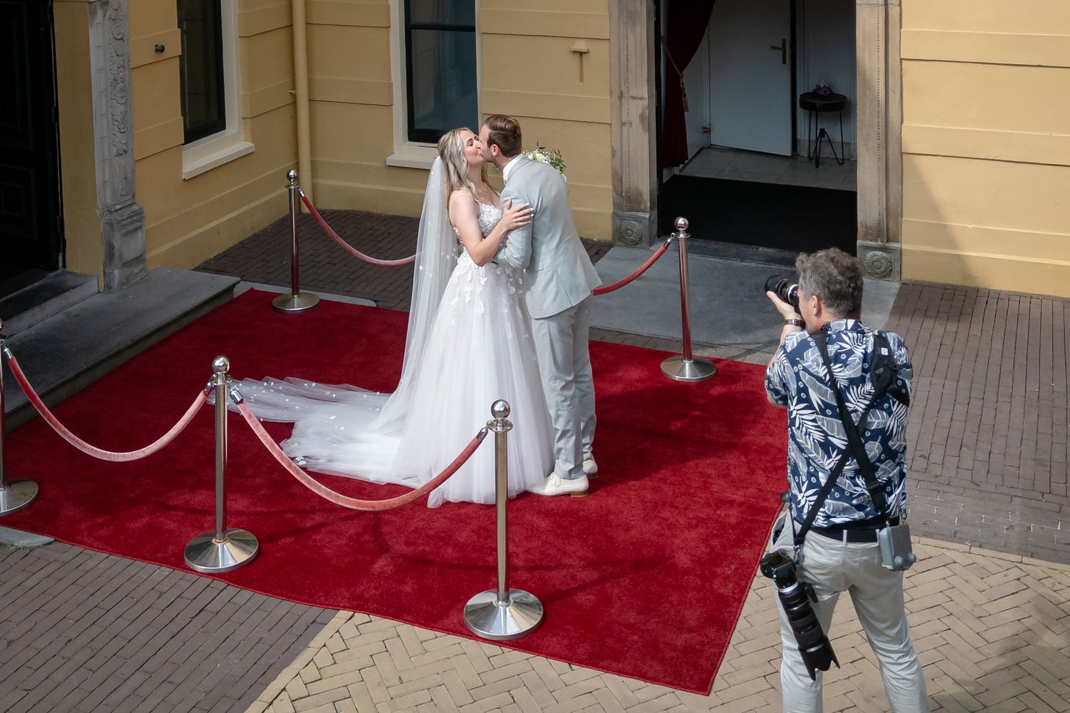 Trouwfotograaf in actie bij first look-first kiss bij bruidspaar op een rode loper met fluwelen afzetkoord bij een klassiek zachtgeel gebouw