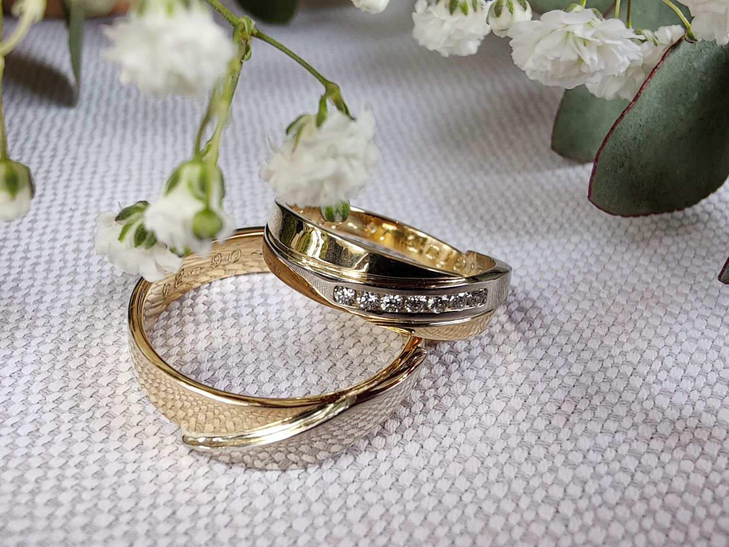 Two gold wedding bands with diamond accents resting on white fabric with flowers.