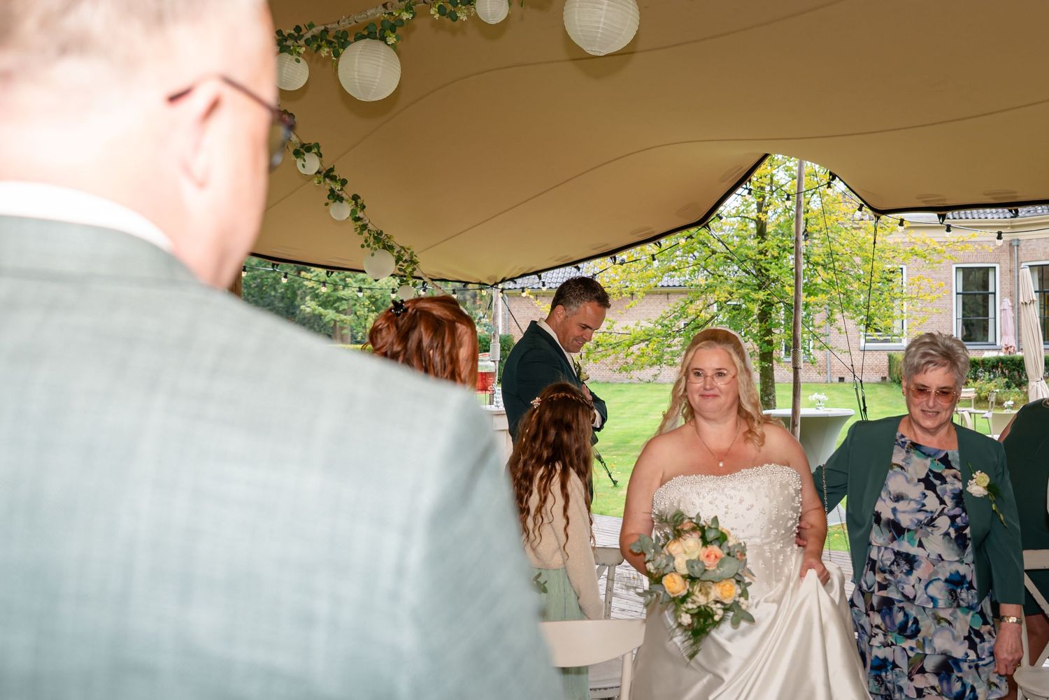 In de stretchtent wordt de bruid door haar moeder aan de bruidegom gegeven. Op deze trouwfoto kijken we over de schouder van de bruidegom.