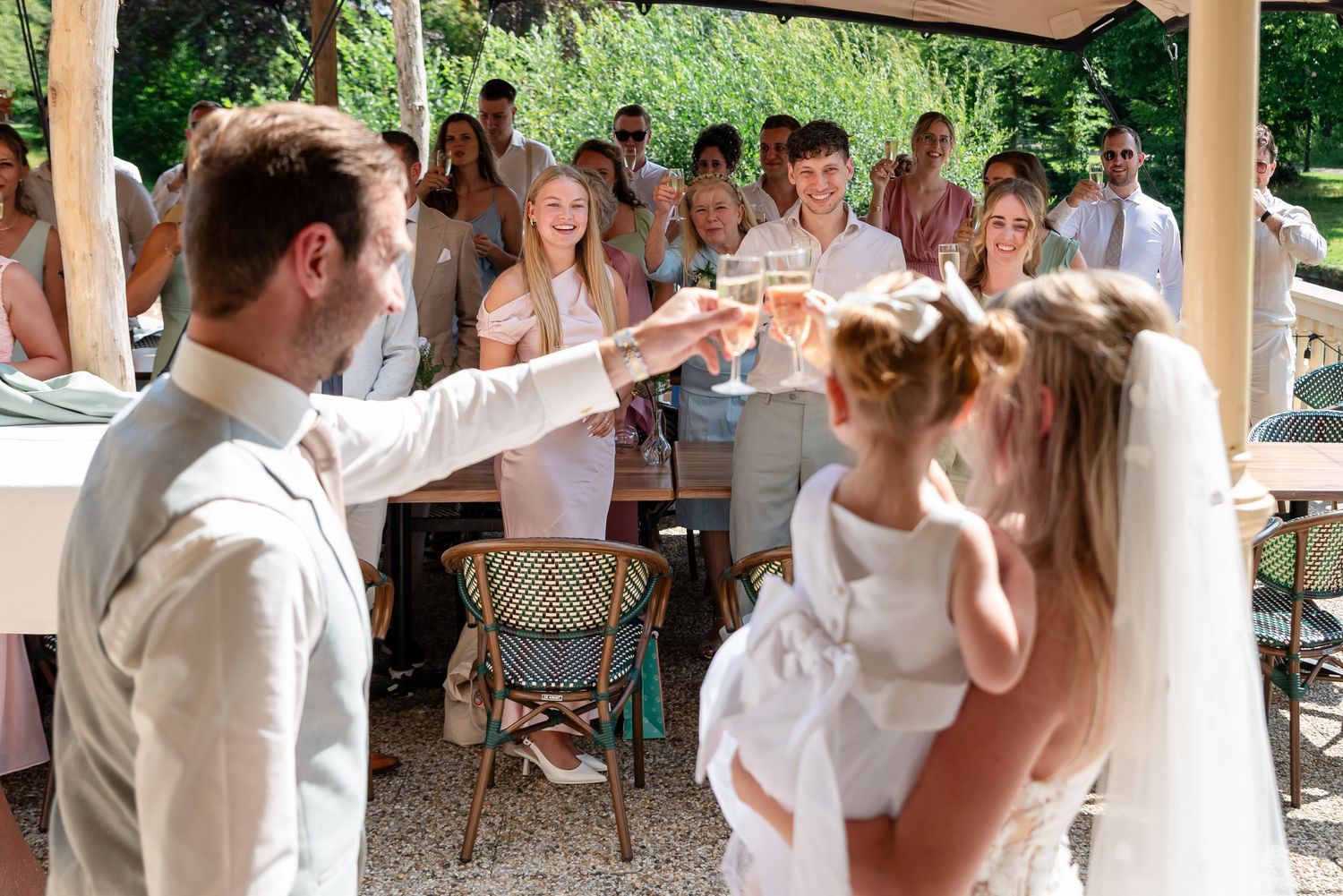 Bride and groom toasting champagne glasses at outdoor wedding reception with guests celebrating in background.