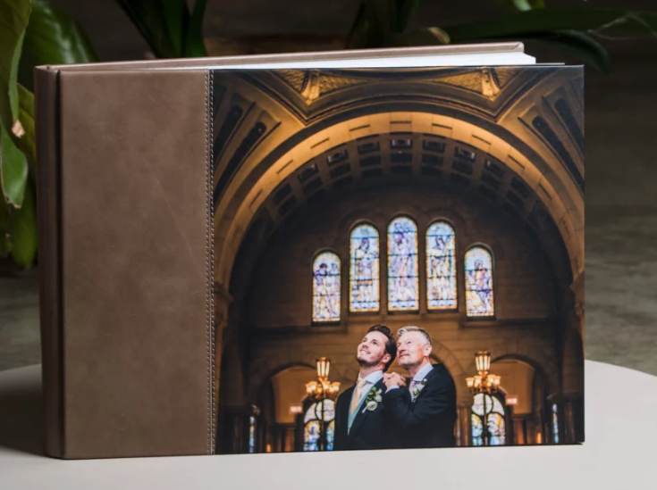 Elegant wedding photo album with leather cover featuring two grooms posing in a stunning cathedral with stained glass windows.