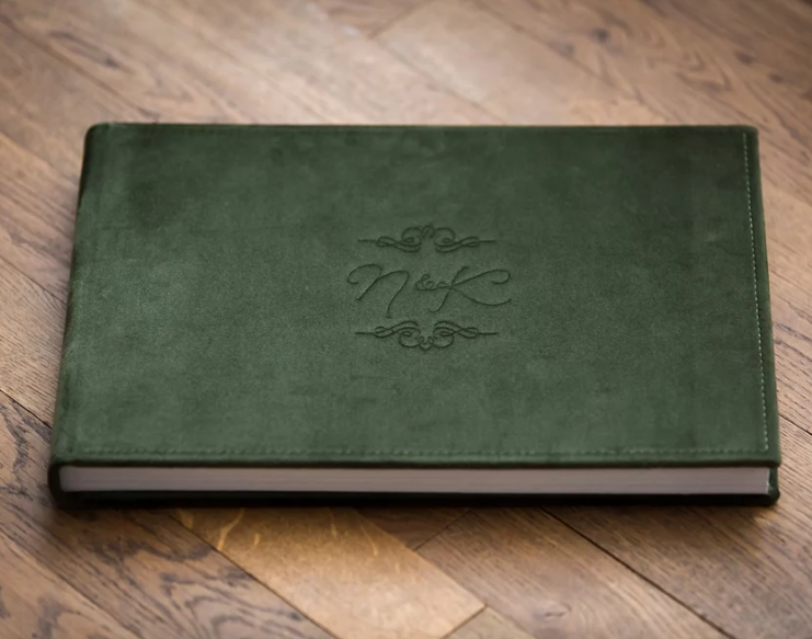 Green velvet embossed monogram guest book lying on rustic wooden surface, ideal for weddings or special events.