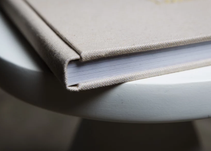 Close-up of a hardcover book with gray linen cover and white pages resting on a round white table.