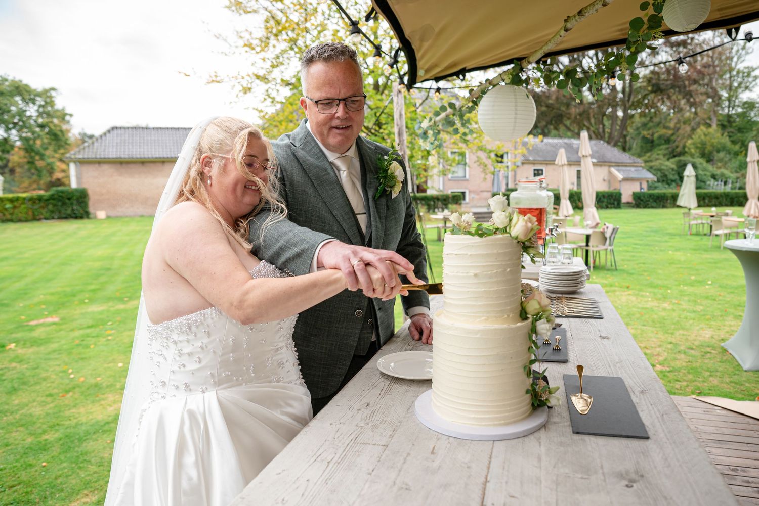 Bride and groom cutting their white tiered wedding cake outdoors on a wooden table in a garden setting.