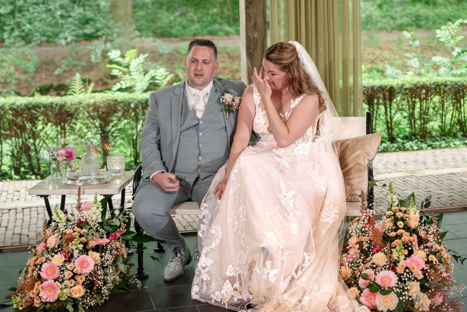Bride and groom seated during outdoor wedding ceremony, bride in lace gown wiping tears, surrounded by pink floral arrangements.