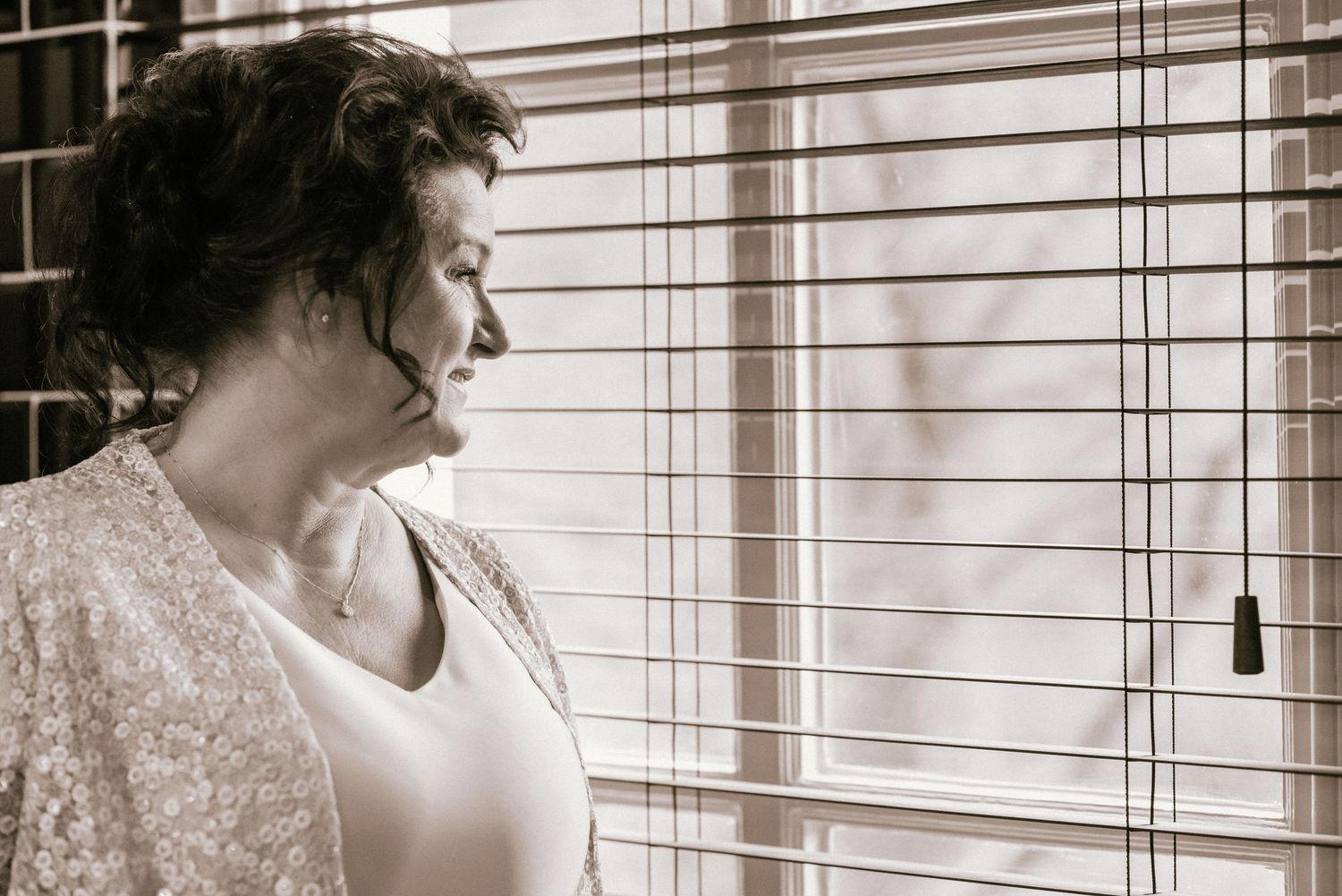 A woman in a lace cardigan gazes thoughtfully through venetian blinds in a sepia-toned portrait.