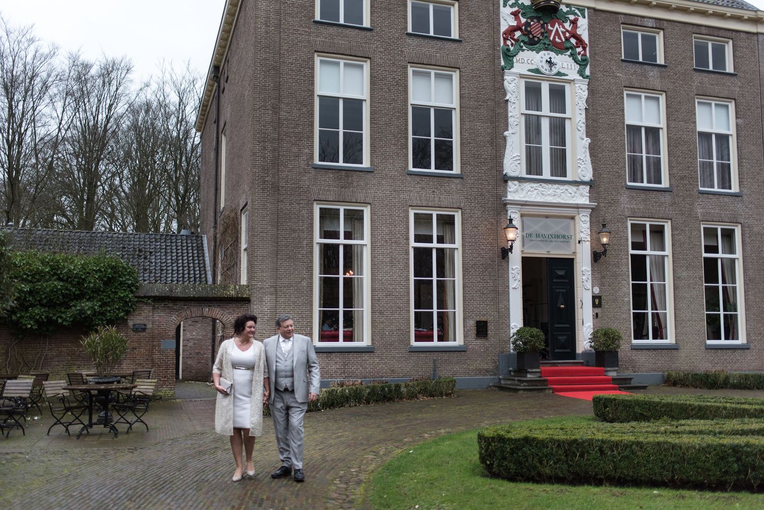 Couple in formal attire standing outside a historic Dutch manor house with red carpet entrance and festive decorations.