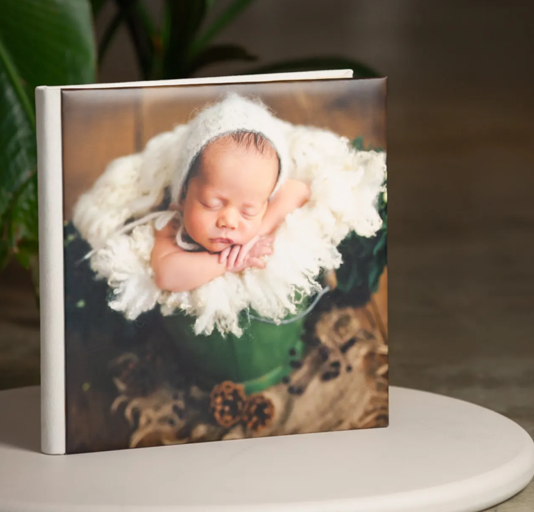 Square photo book with a newborn baby sleeping in a green bucket with white fur, displayed on a white surface.
