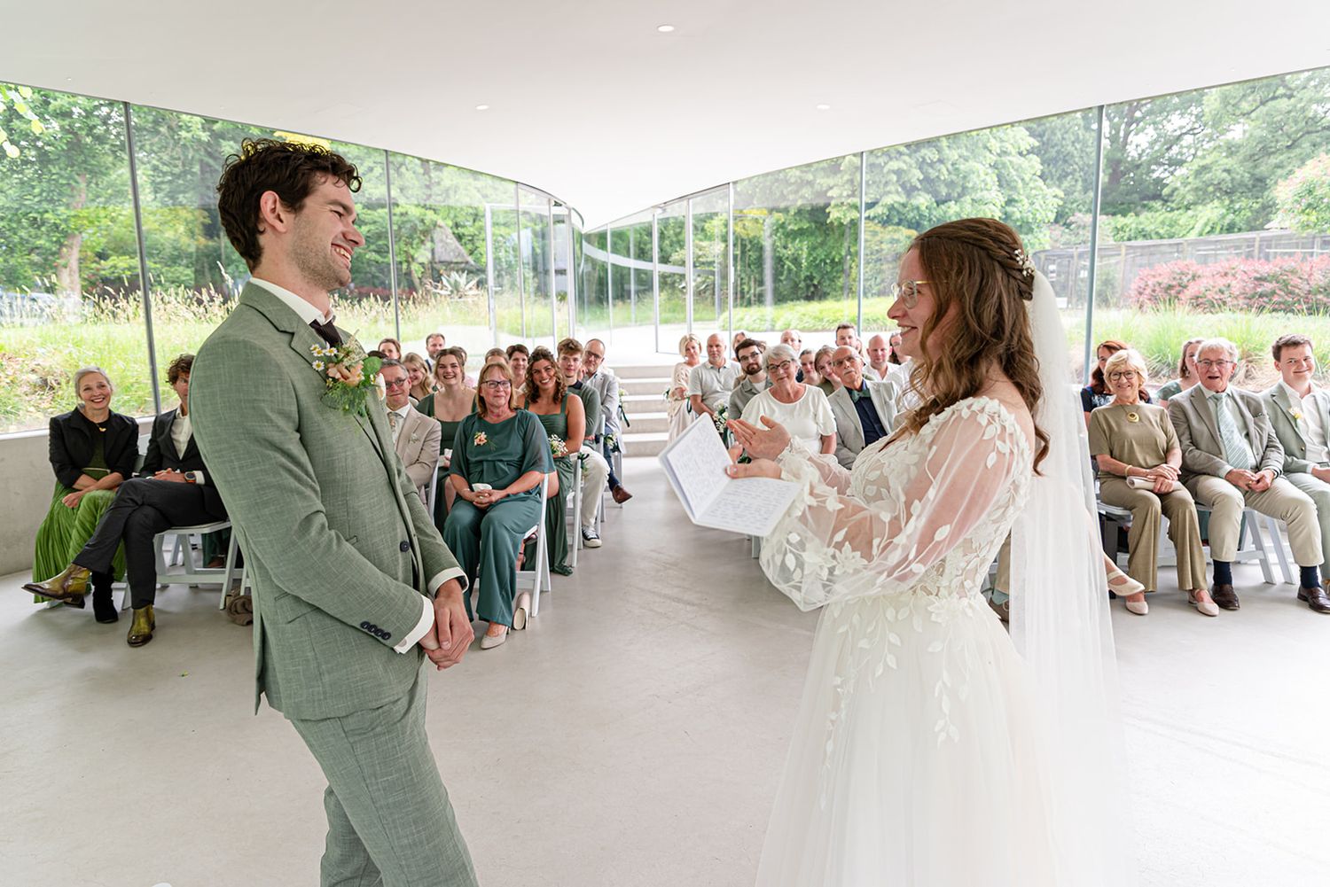 Bride reads vows to groom in elegant glass wedding venue with seated guests in background.
