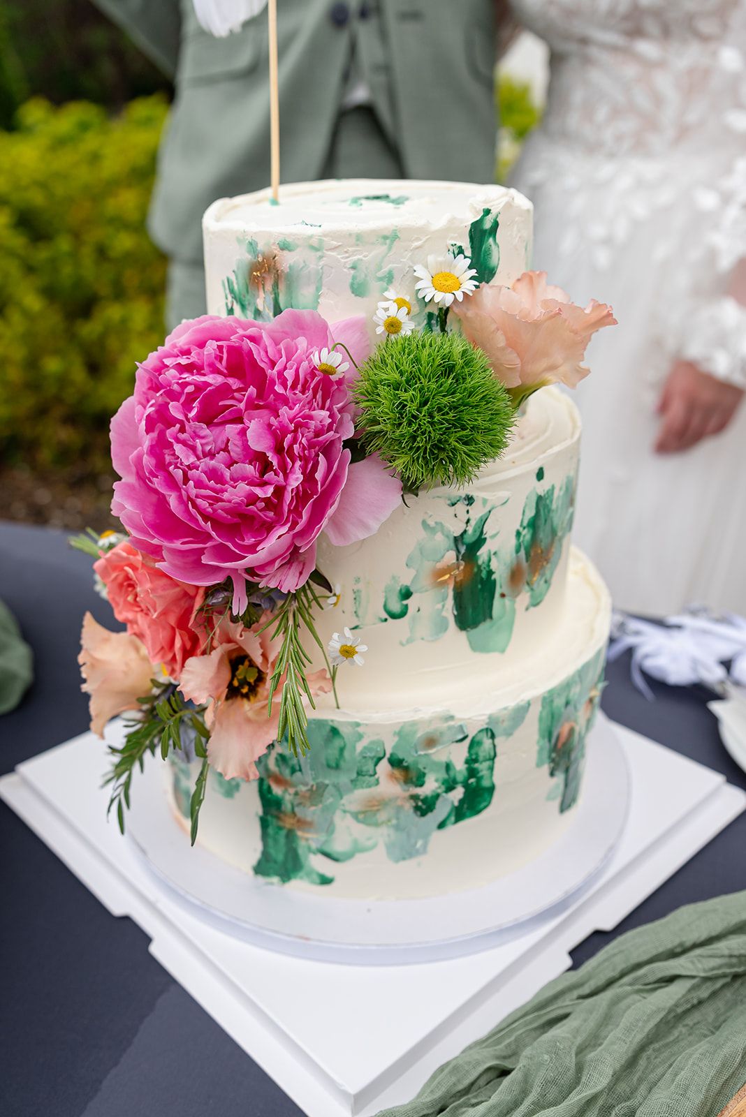 Two-tier white wedding cake with green paint strokes and fresh pink peonies, daisies, and coral flowers outdoors.