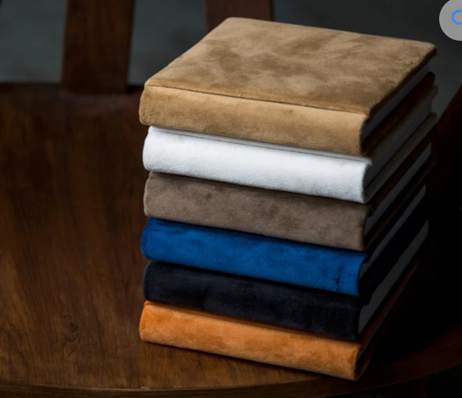 Stack of seven suede notebooks in tan, white, gray, brown, blue, black, and orange on a wooden table.