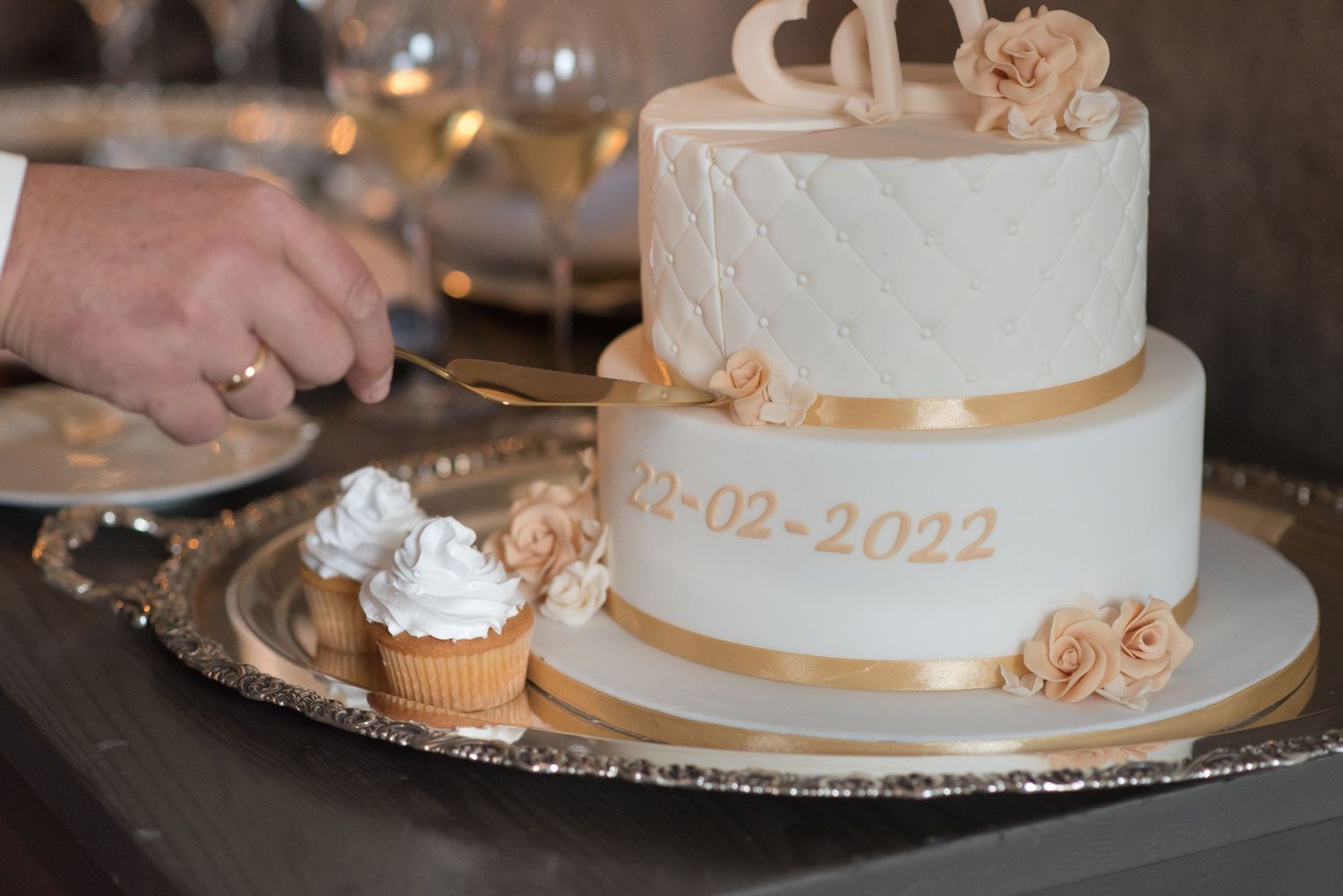 Two-tier white and gold wedding cake with date 22-02-2022, roses, and a cupcake on a silver tray being cut.