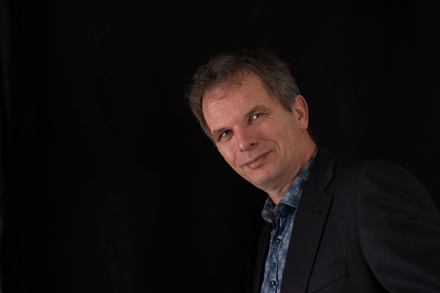 Middle-aged man in a dark blazer smiling against a black background in a professional portrait photo.