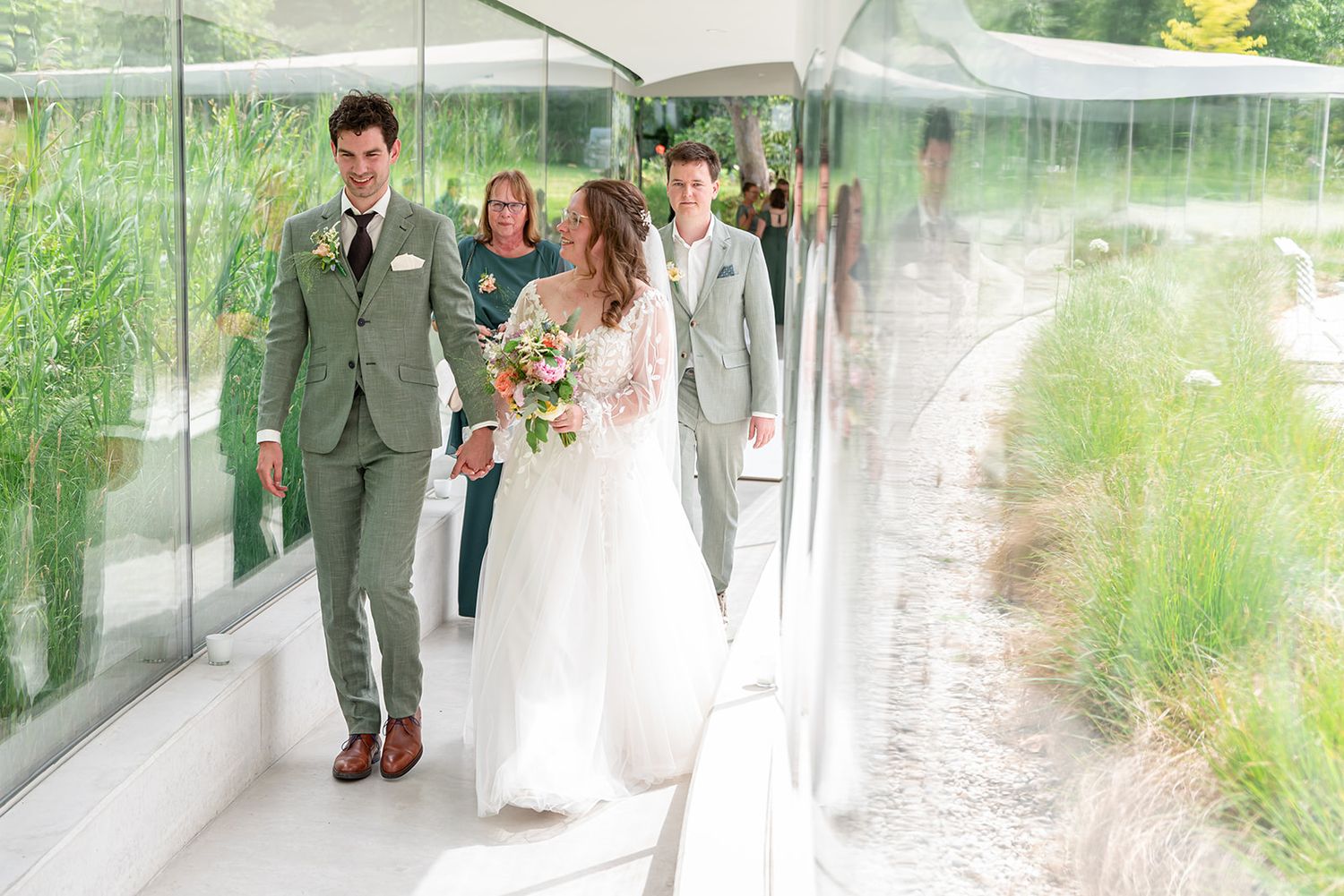 Bride and groom walking through a modern glass corridor with wedding guests, surrounded by lush greenery.