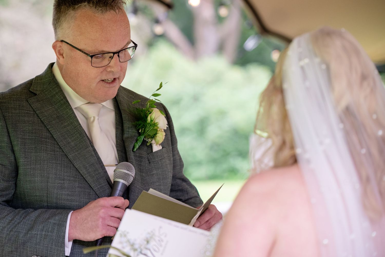 Bruidegom leest in een groen trouwpak en een microfoon in de hand gelofte voor aan zijn bruid. We kijken mee over de schouder van de bruid.