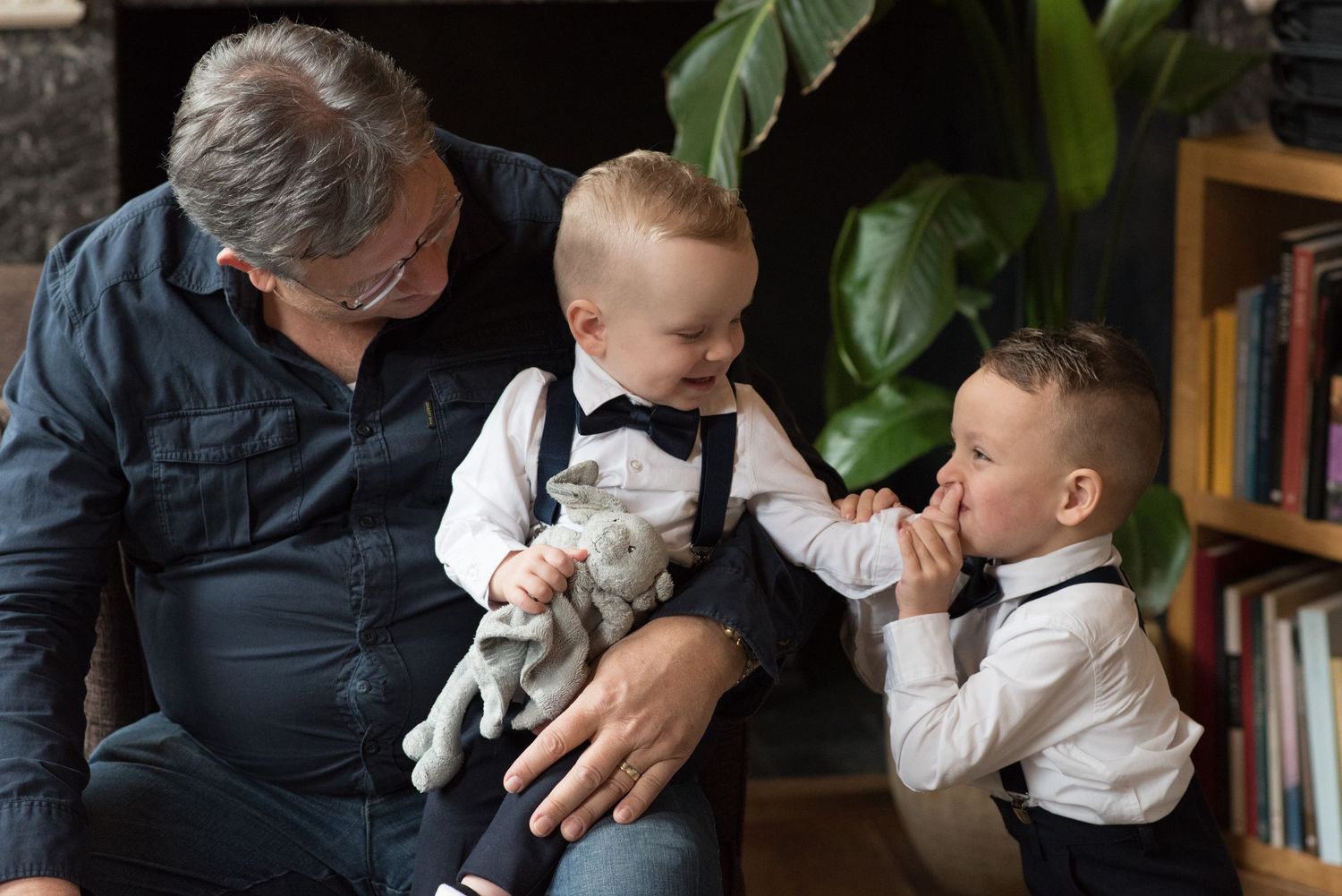 A grandfather smiles warmly while holding two young boys dressed in suspenders and bow ties in a cozy home setting.