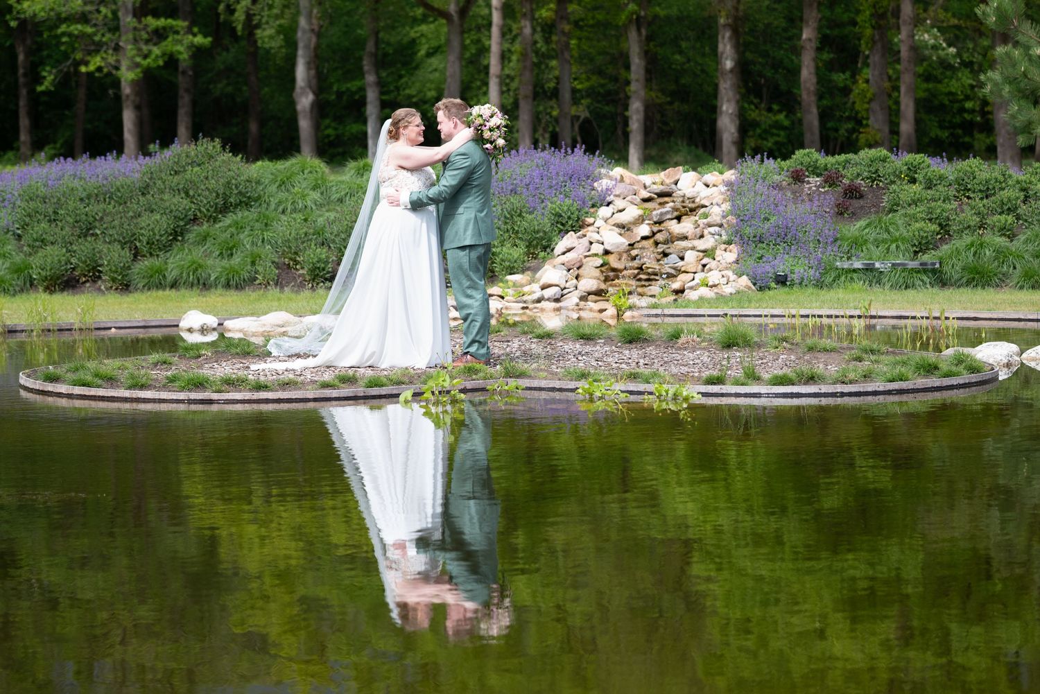 Bride and groom kissing beside a tranquil pond with their reflection visible in lush garden.