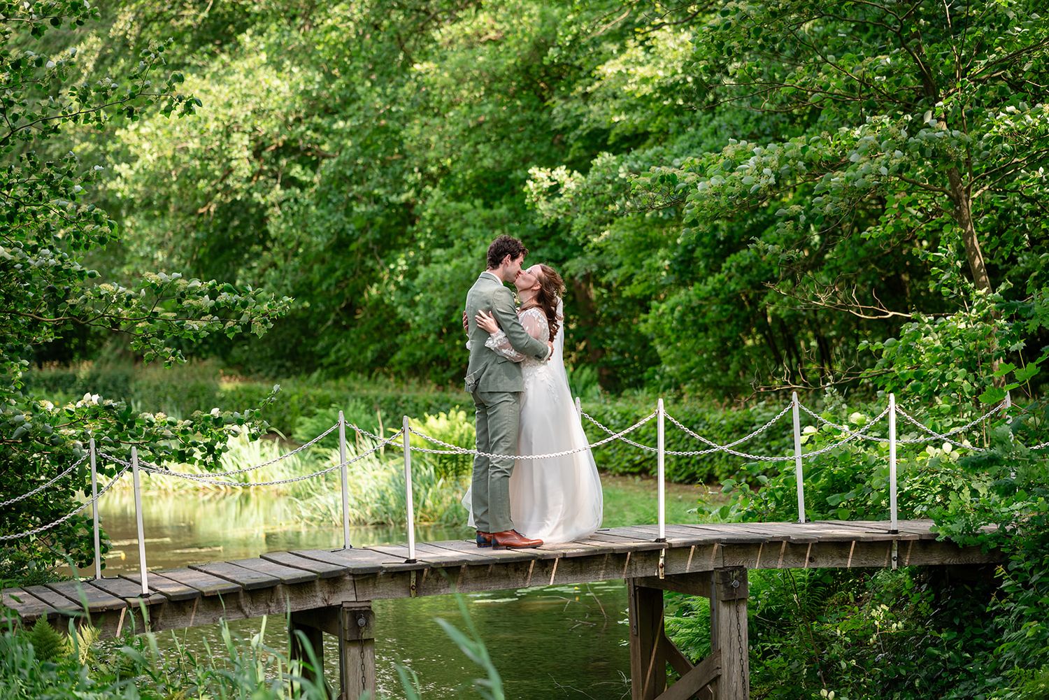 Couple sharing a romantic kiss on a wooden bridge surrounded by lush green trees near a tranquil pond.