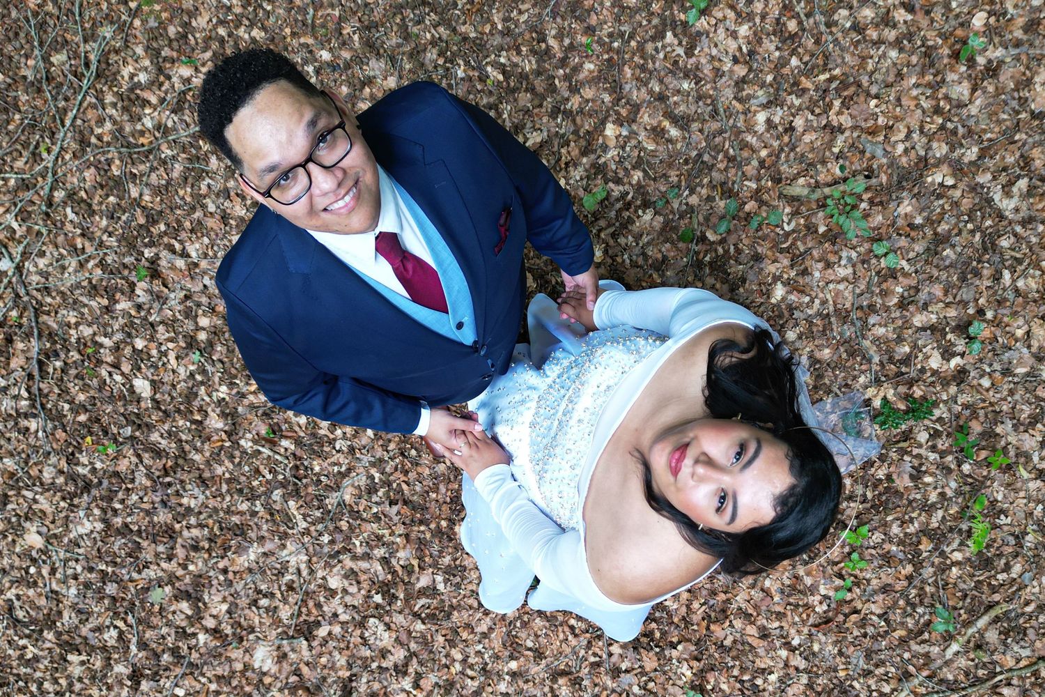 Aerial view of a couple in wedding attire holding hands on a bed of autumn leaves, smiling up at the camera.