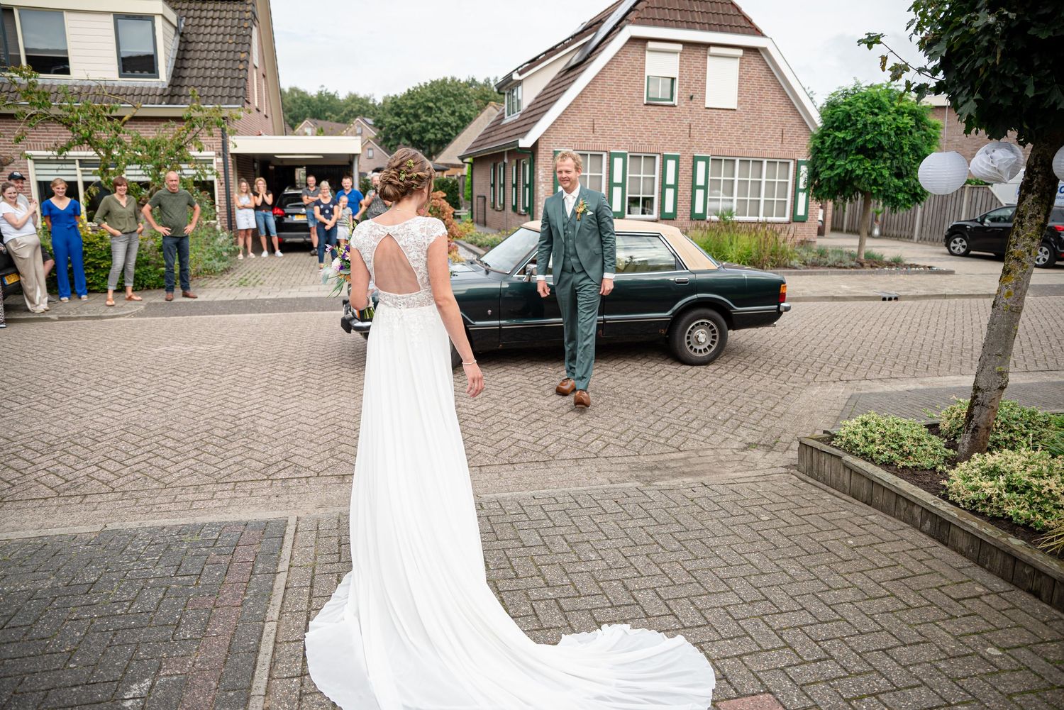 Bride in white gown with train stands facing groom in teal suit near vintage black car on brick driveway at wedding.