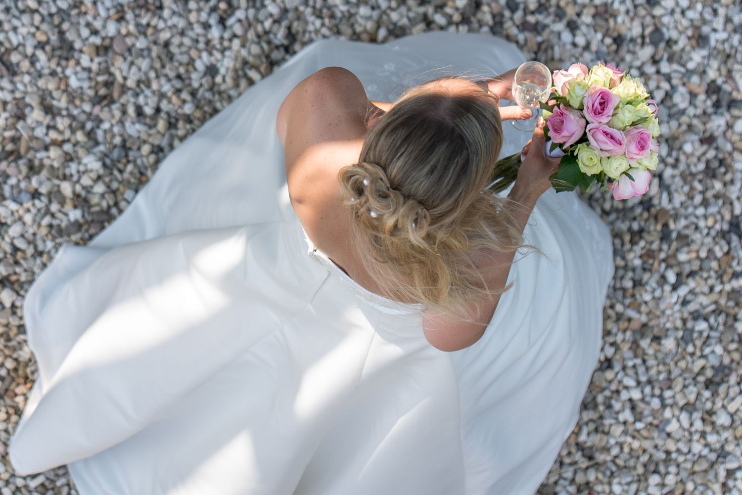 Aerial view of a bride in a white gown sitting on pebbles, holding a pink and white bouquet.