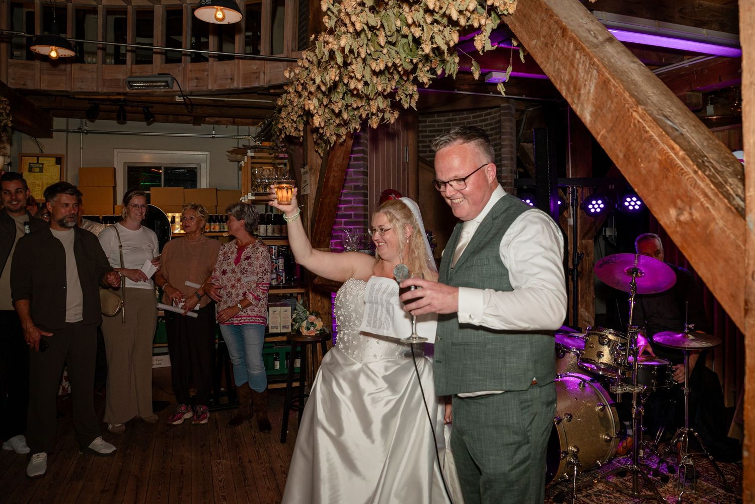 Bride and groom share first dance in rustic barn venue with guests watching and drum kit visible in background.
