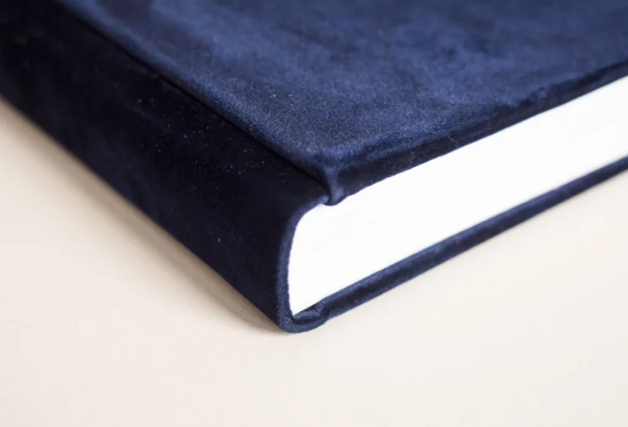 Close-up of a hardcover book with a luxurious navy blue velvet cover and white pages on a beige surface.
