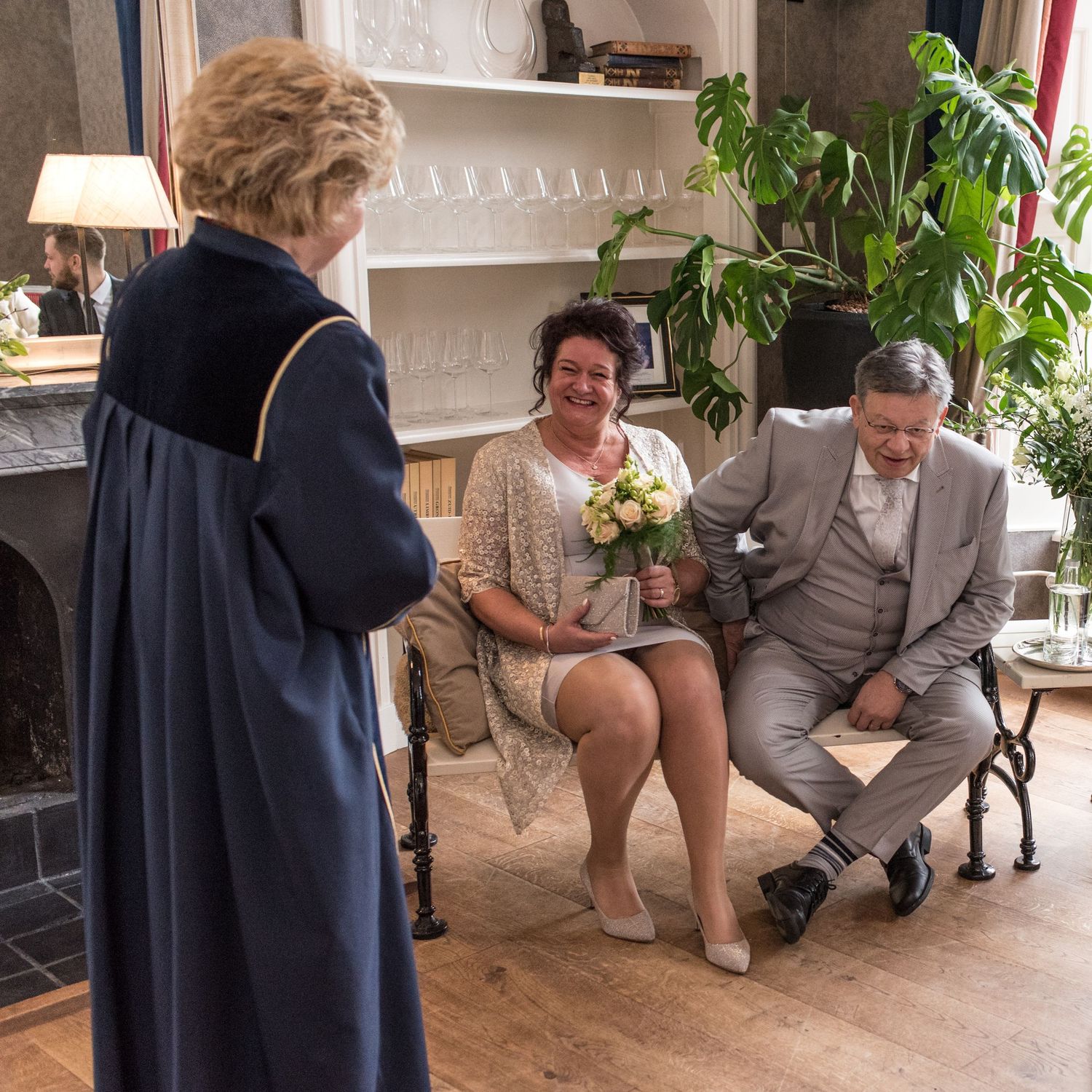 Elderly couple sits with bouquet during intimate wedding ceremony officiated by robed official in elegant room.
