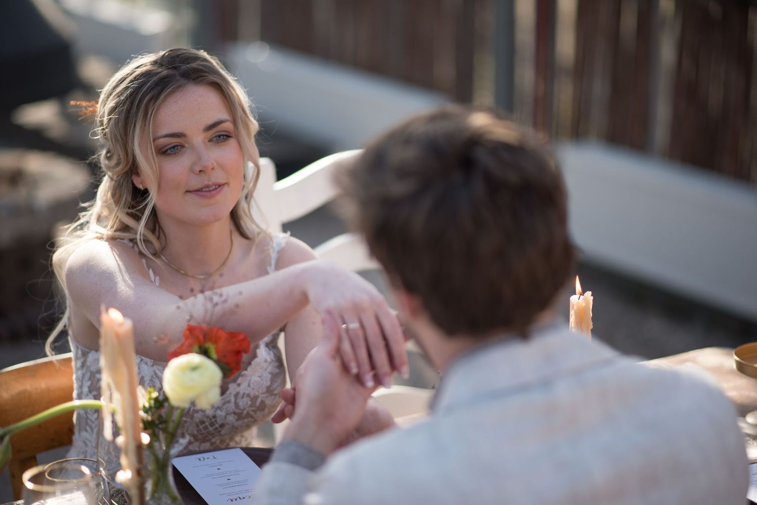 A blonde bride in a lace gown holds hands with her partner at a romantic candlelit outdoor table.