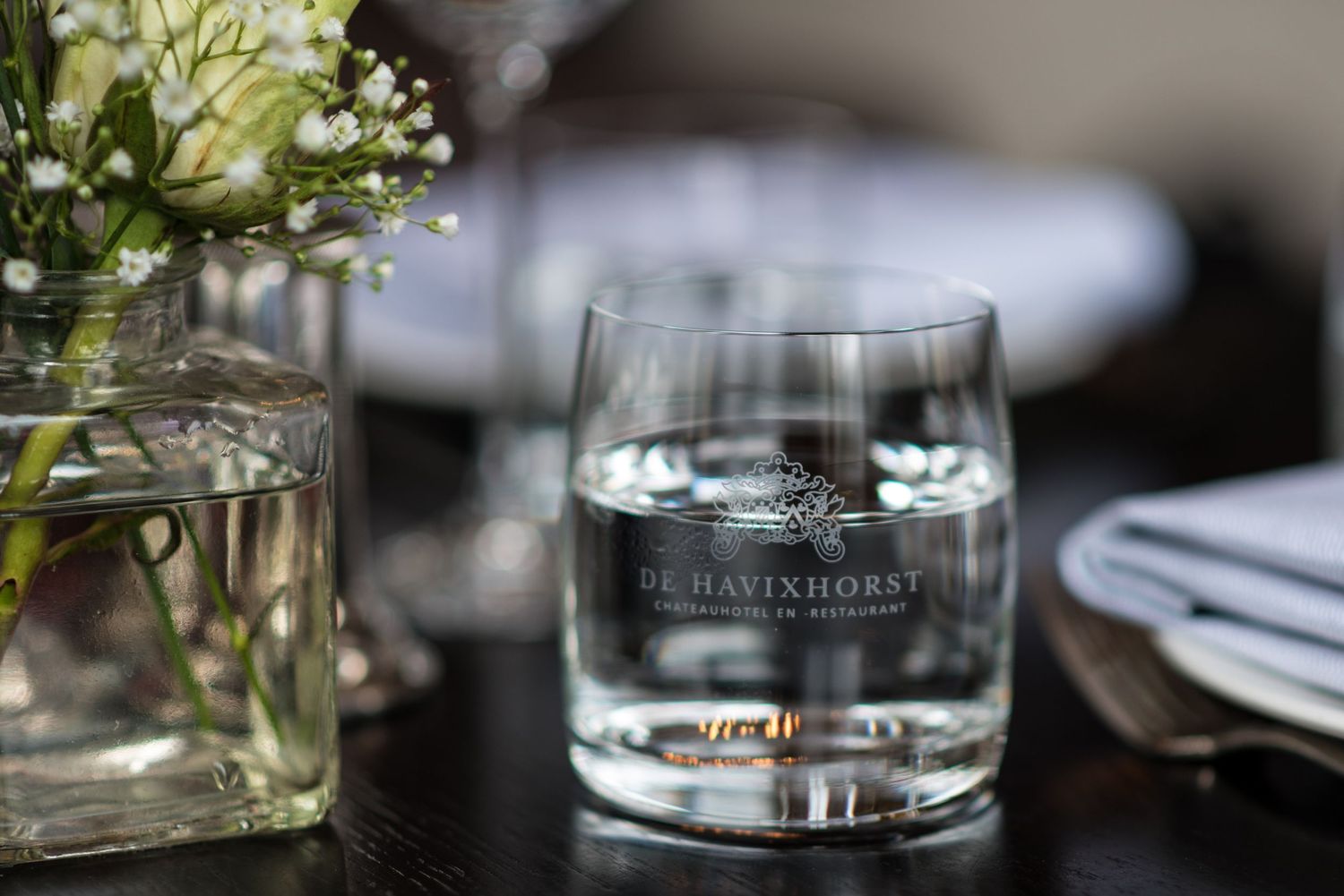 De Havinhorst branded glass tumbler with water on a dark table beside a floral arrangement and folded napkins.