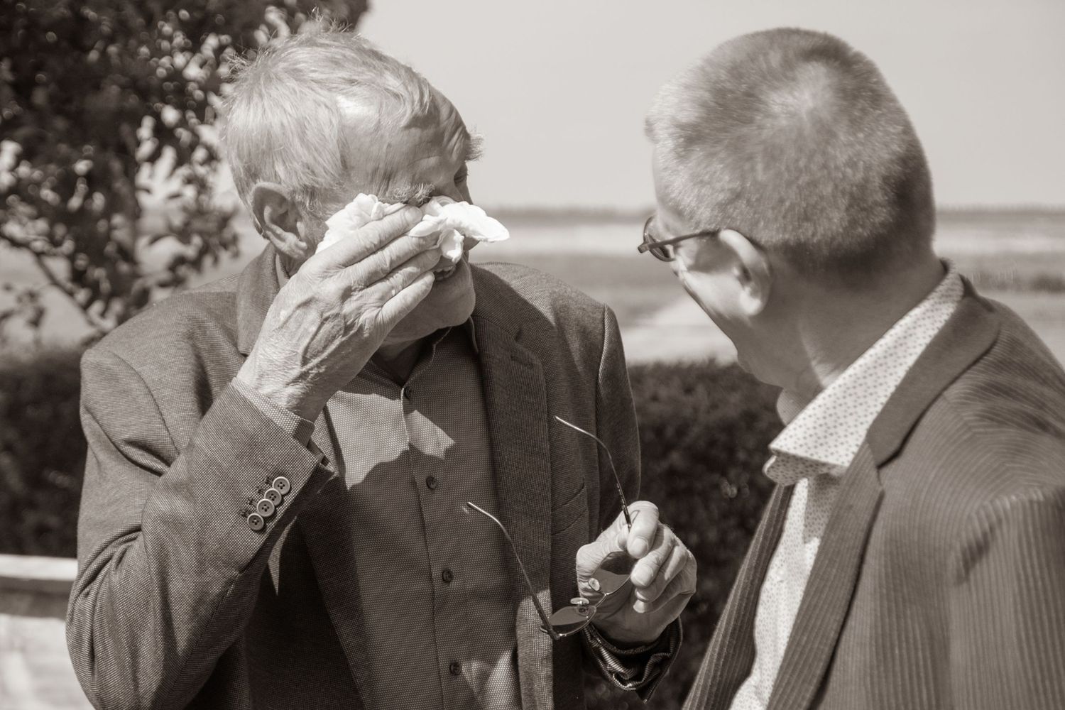 Twee bruiloftsgasten in gesprek. De oude man is erg emotioneel. Hij houdt zijn bril vast terwijl hij met een zakdoek in de ogen wrijft.