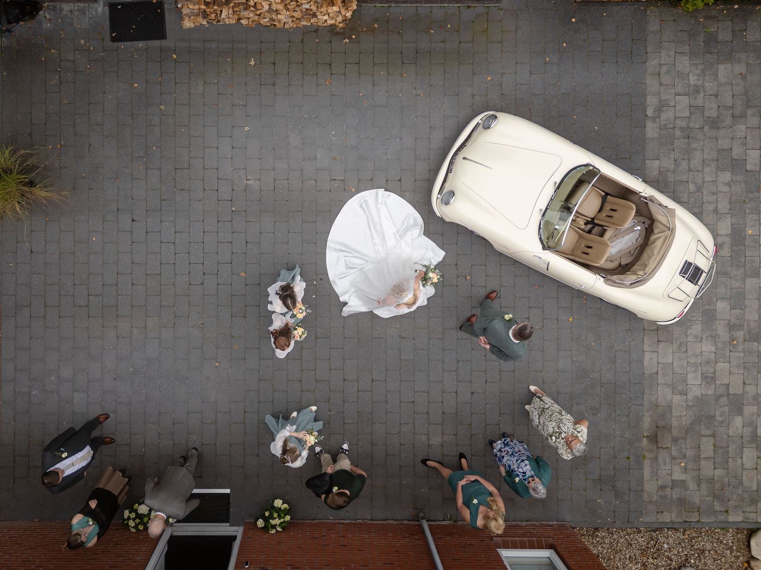Droneopname van een bruidspaar met een witte Porsche 356 cabrio als trouwauto, op de oprit van hun huis. De sleep van de trouwjurk ligt mooi