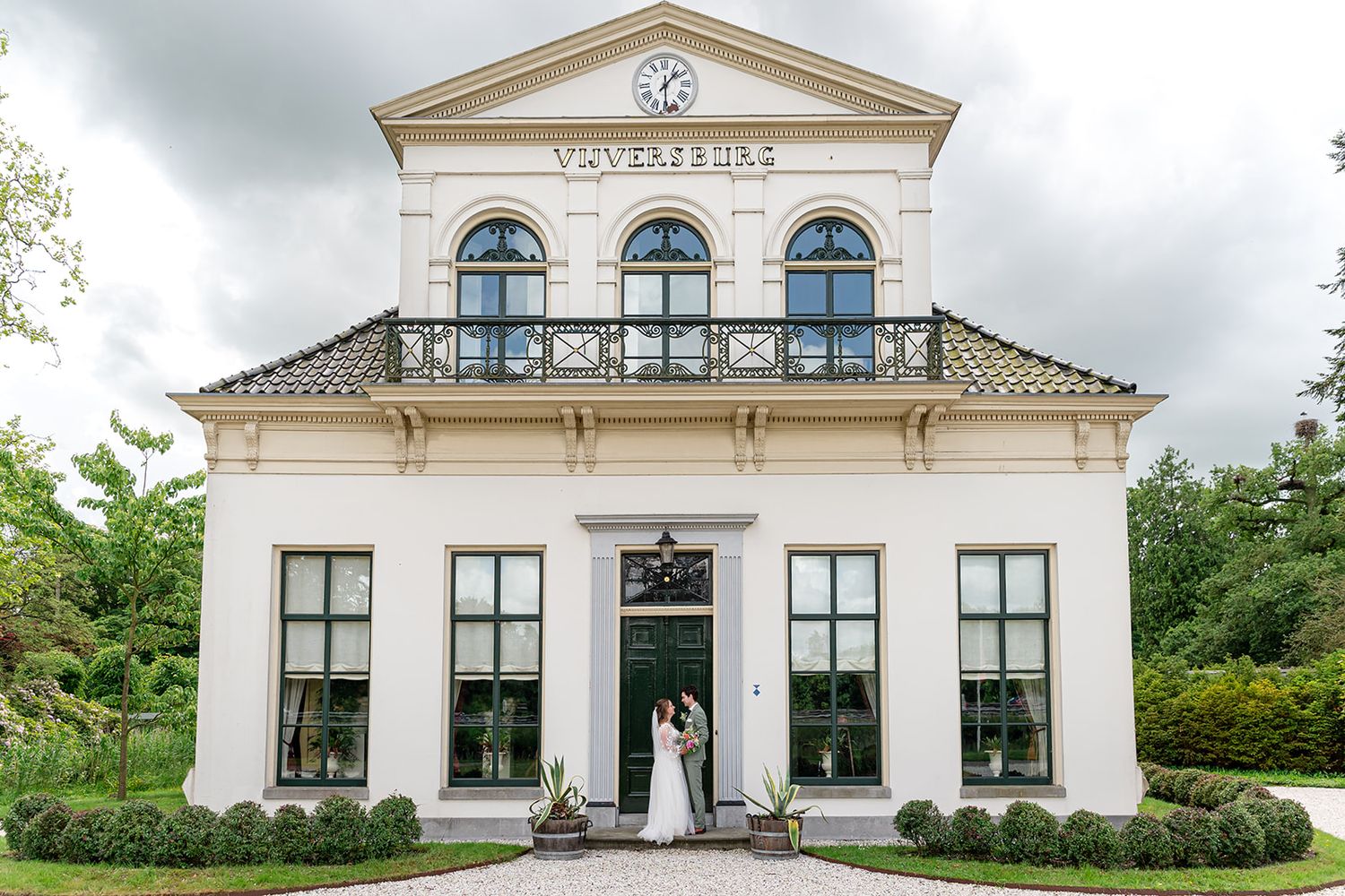 Bride and groom posing at entrance of elegant white neoclassical wedding venue with clock tower and balcony.
