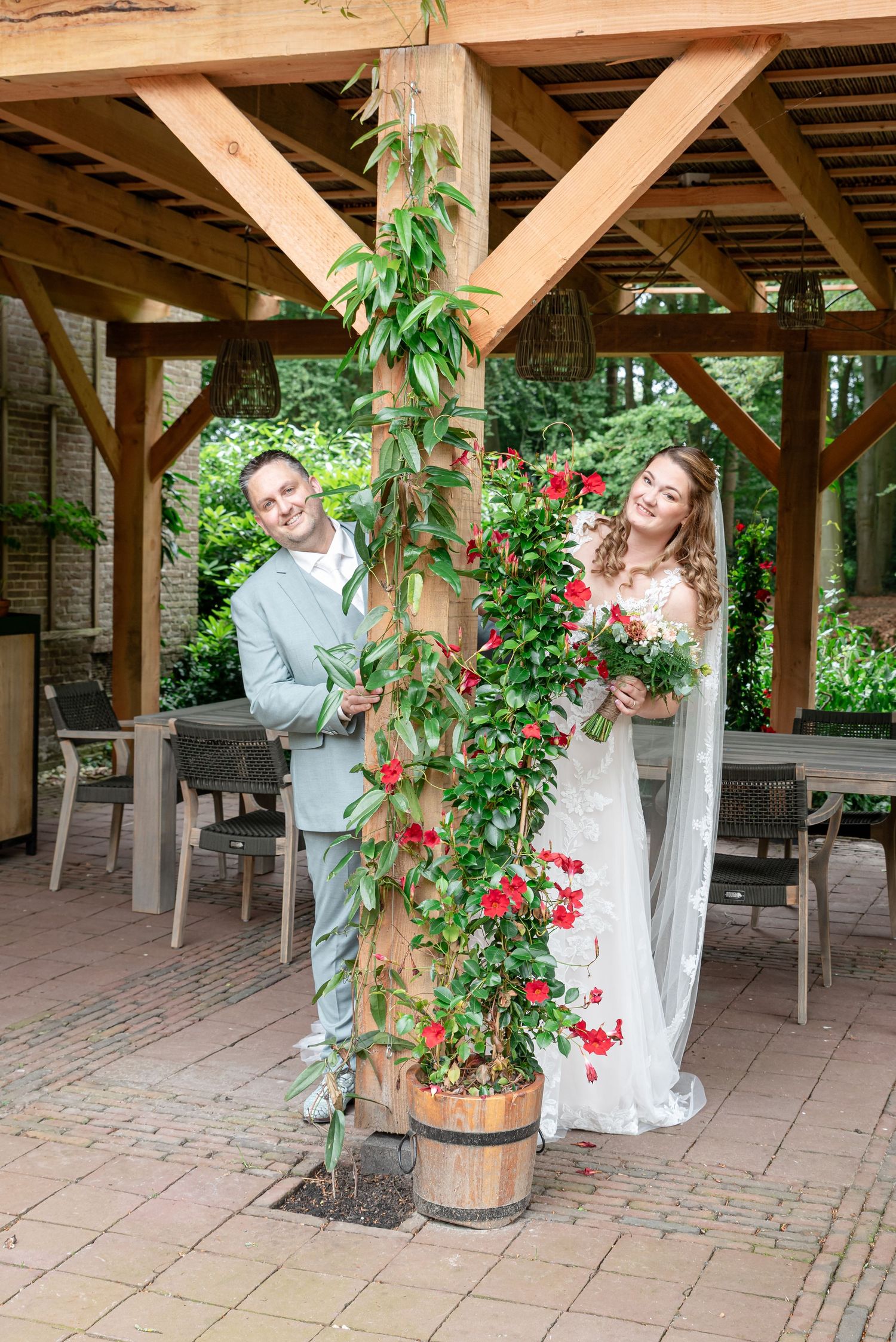 Bride and groom posing beside a flowering red vine on a wooden pillar under a pergola.