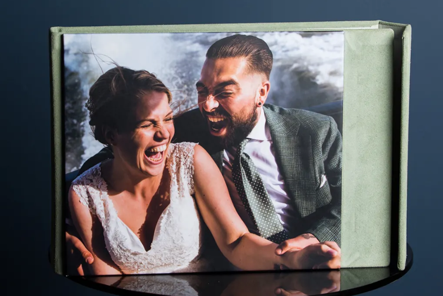 Laughing bride in a white lace dress and groom in a green suit embrace in a wedding photo displayed in a sage green keepsake album.