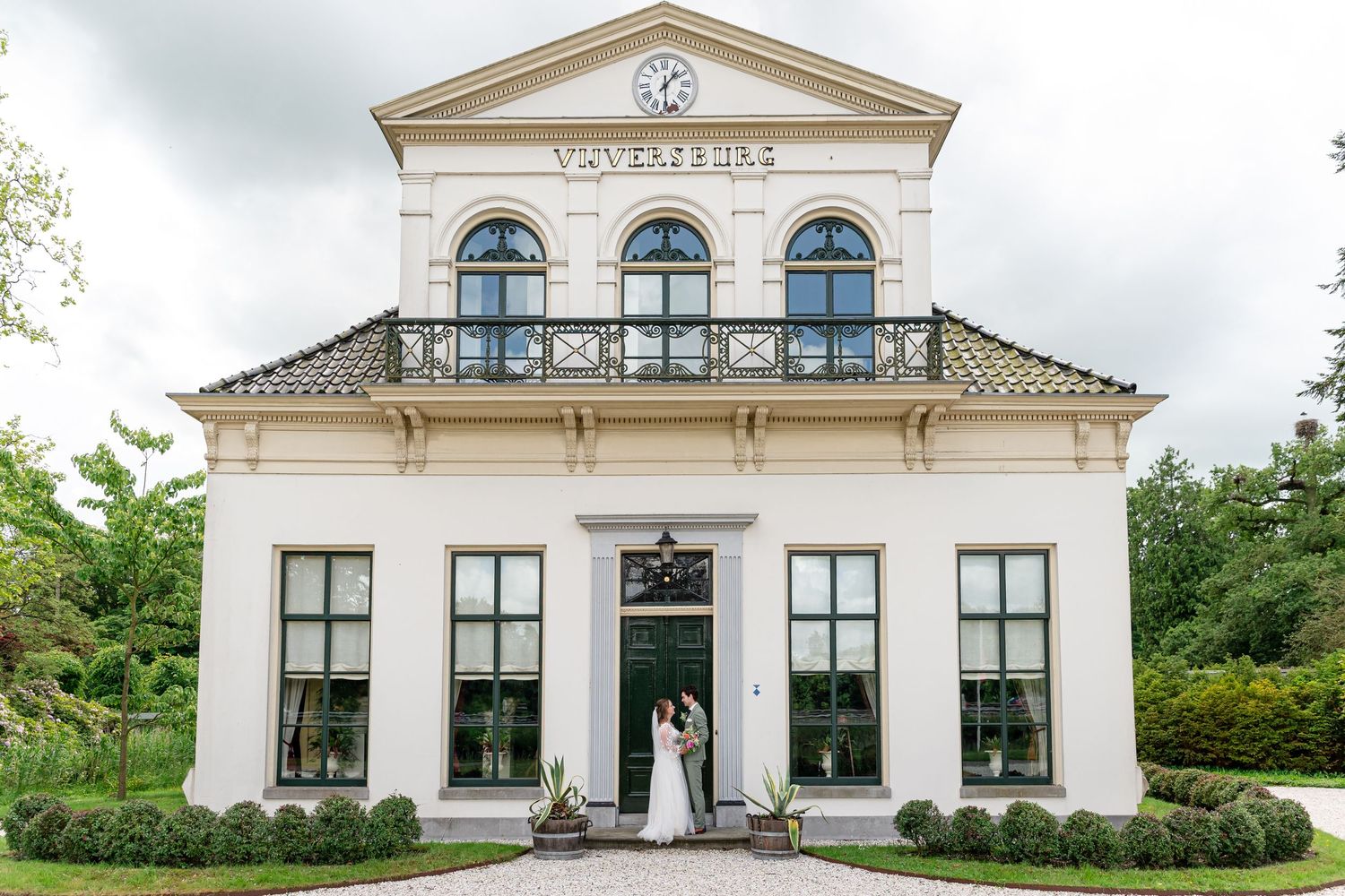 Bride and groom posing in front of elegant white Dutch villa with clock tower.