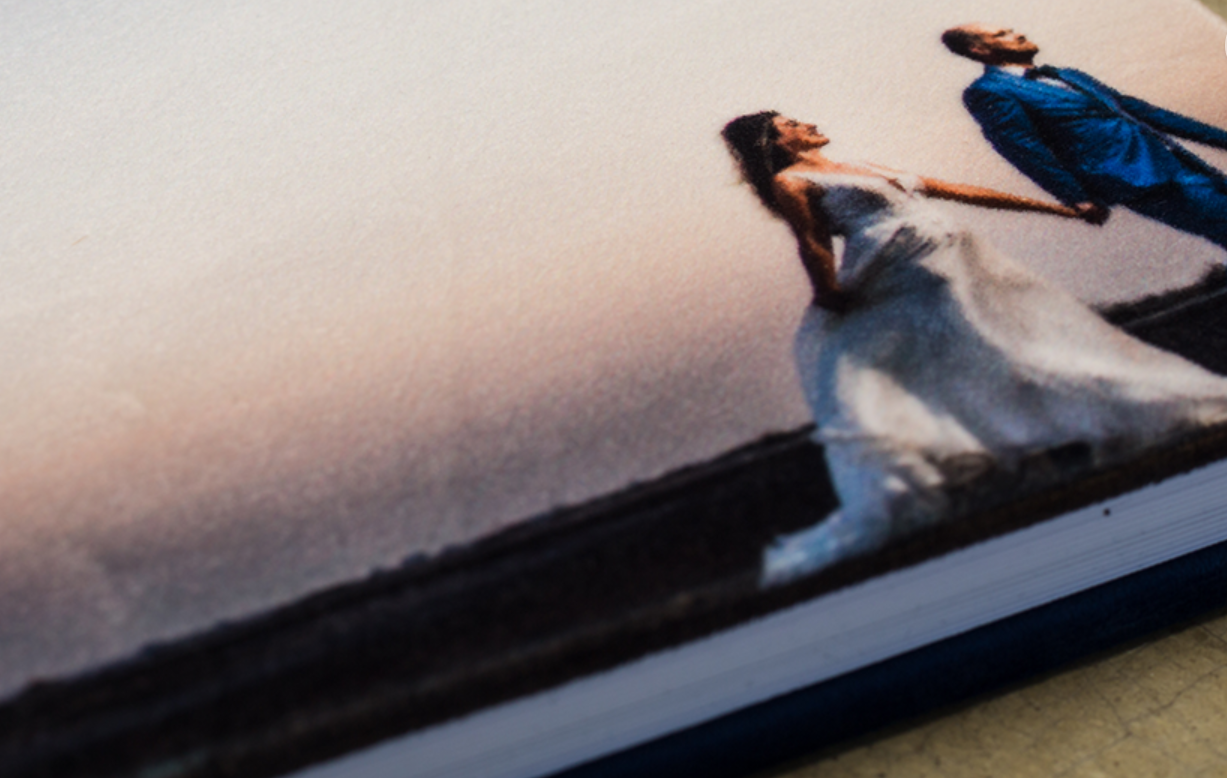 Close-up of a wedding album cover showing a bride in a flowing white gown and a groom in a blue suit walking hand in hand at sunset.