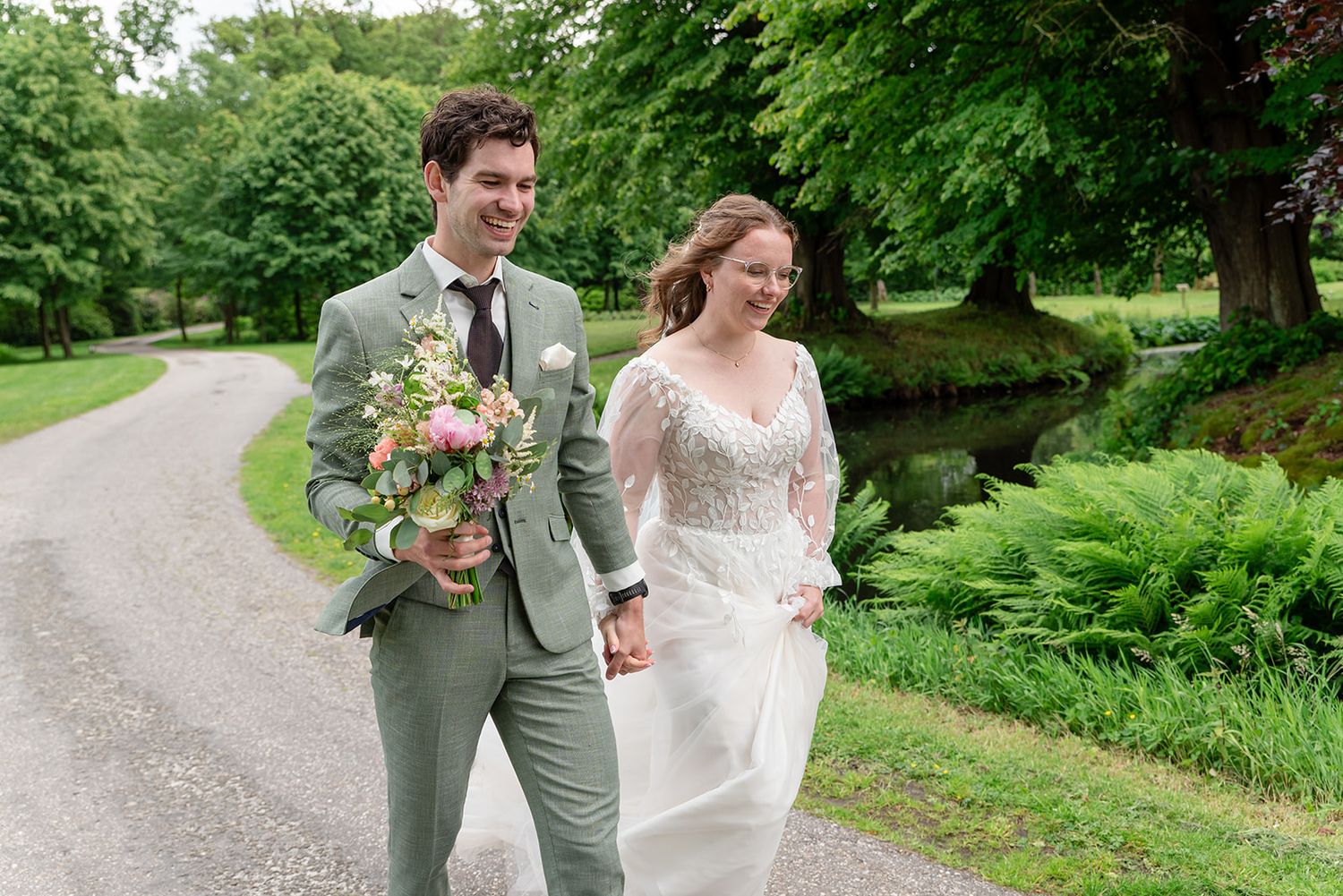 Bride and groom walking hand in hand on a garden path, groom in sage suit holding bouquet, bride in lace gown.