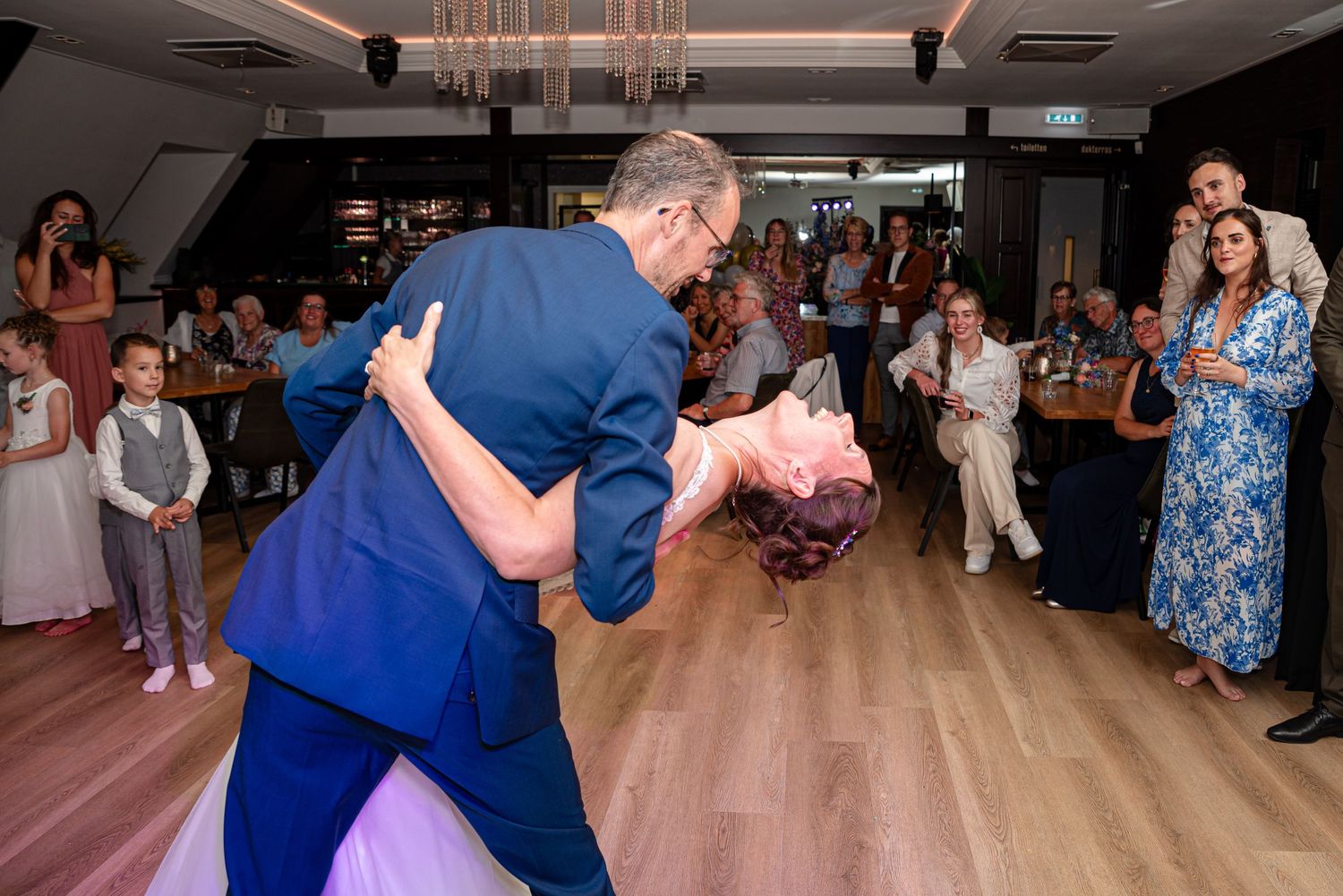 Groom dipping bride during lively first dance at wedding reception with guests watching joyfully.