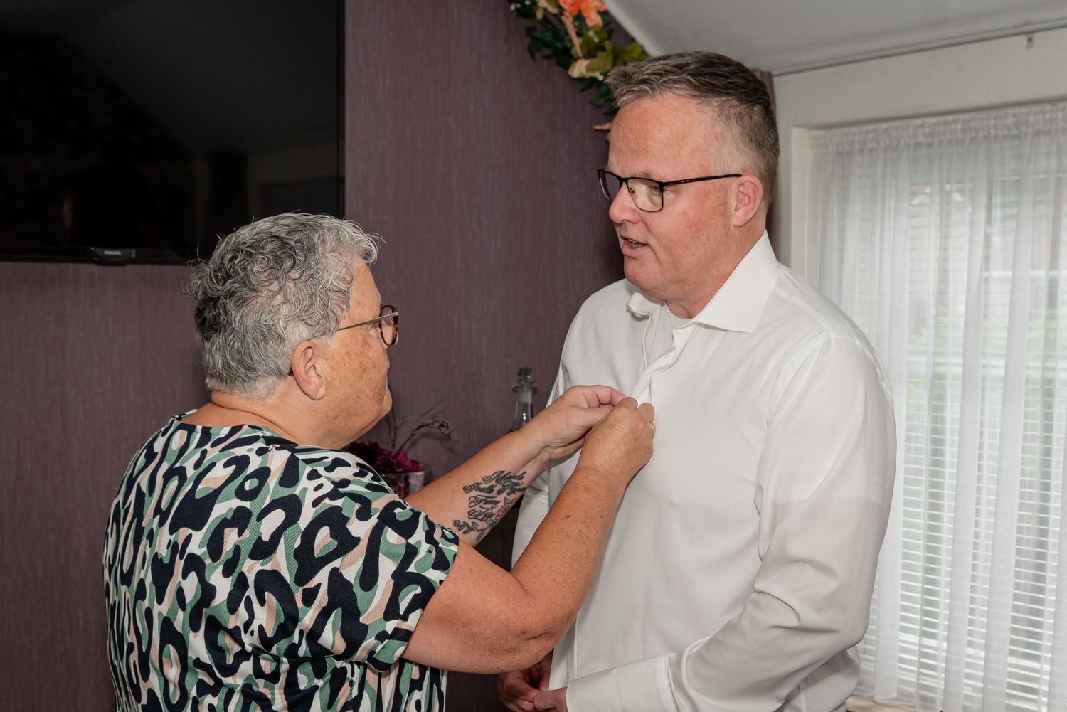 Bruidegom wordt door zijn moeder geholpen bij het aantrekken van zijn bruidspak, hier met het dichtknopen van zijn overhemd.