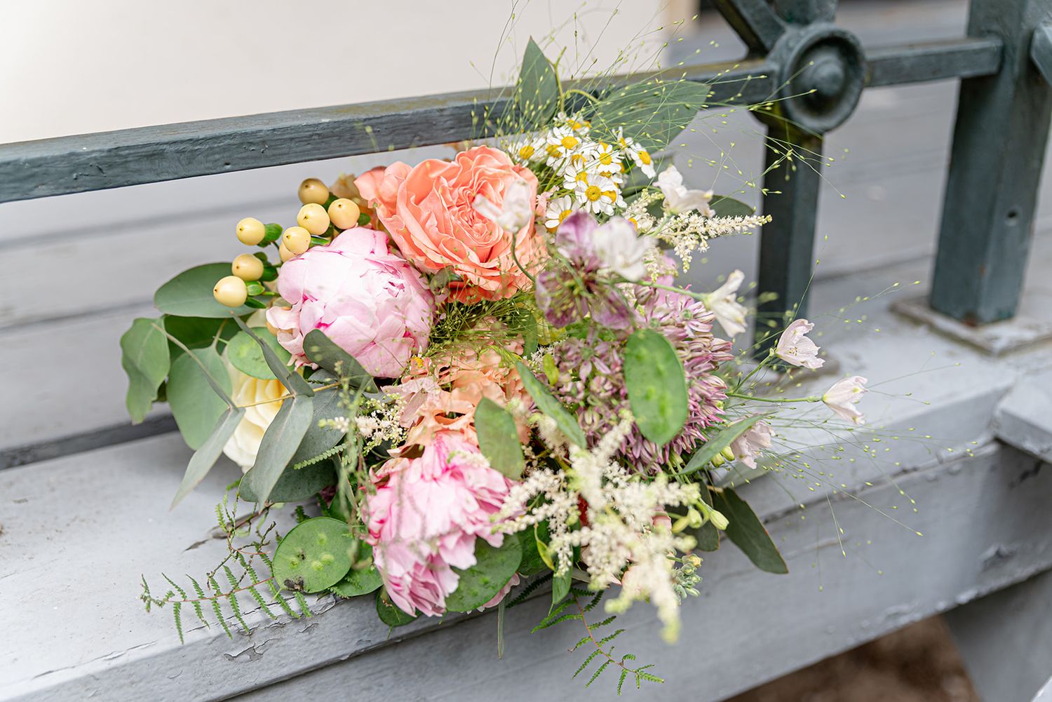 A romantic bridal bouquet with pink peonies, coral roses, berries, and wildflowers resting on a gray bench.