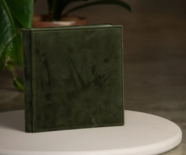Dark green velvet square photo album standing upright on a white table with a plant in the background.