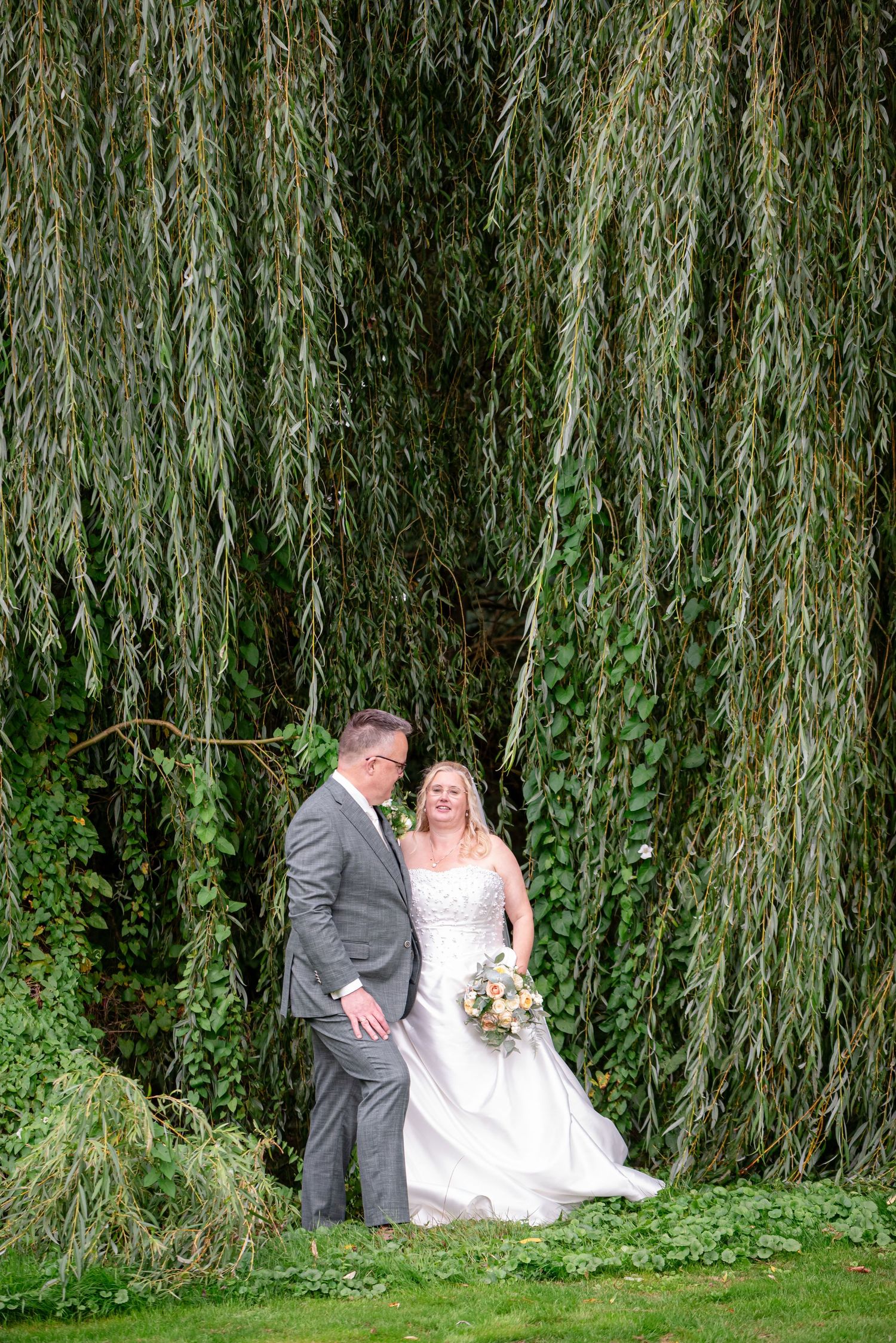 Trouwfoto van Bruid en Bruidegom in een romantische pose voor een prachtige treurwilg, de bruid houdt een bruidsboeket vast.