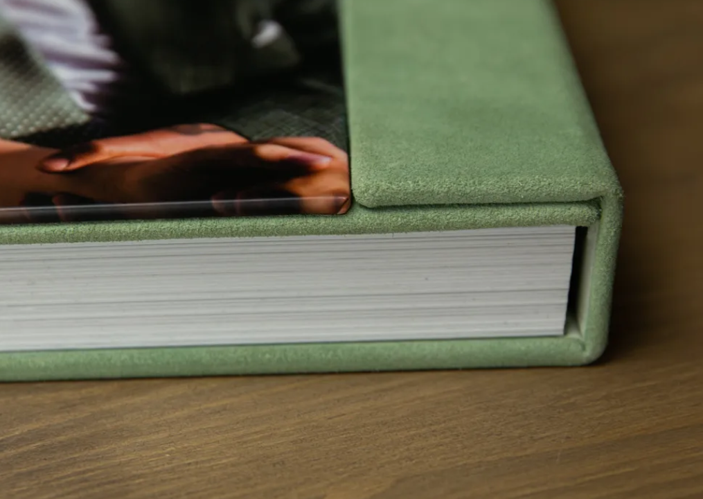 Close-up of a sage green linen-bound photo album showing thick white pages and a printed photo cover on a wooden table.