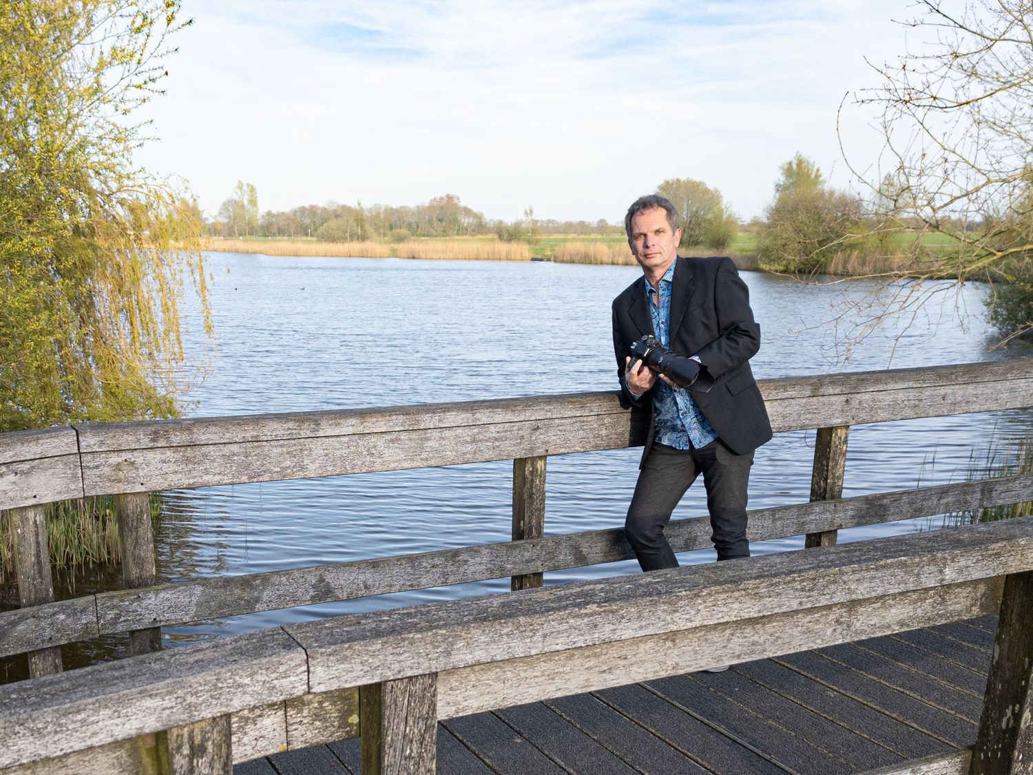 Man with camera standing on wooden bridge overlooking a serene lake surrounded by trees on a clear day.