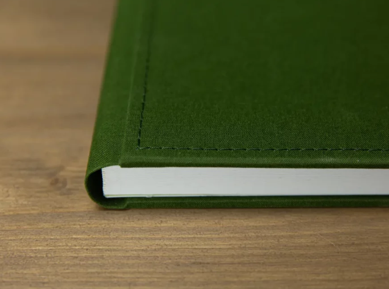 Green hardcover notebook with stitched binding resting on a wooden surface, showcasing durable fabric cover and white pages.