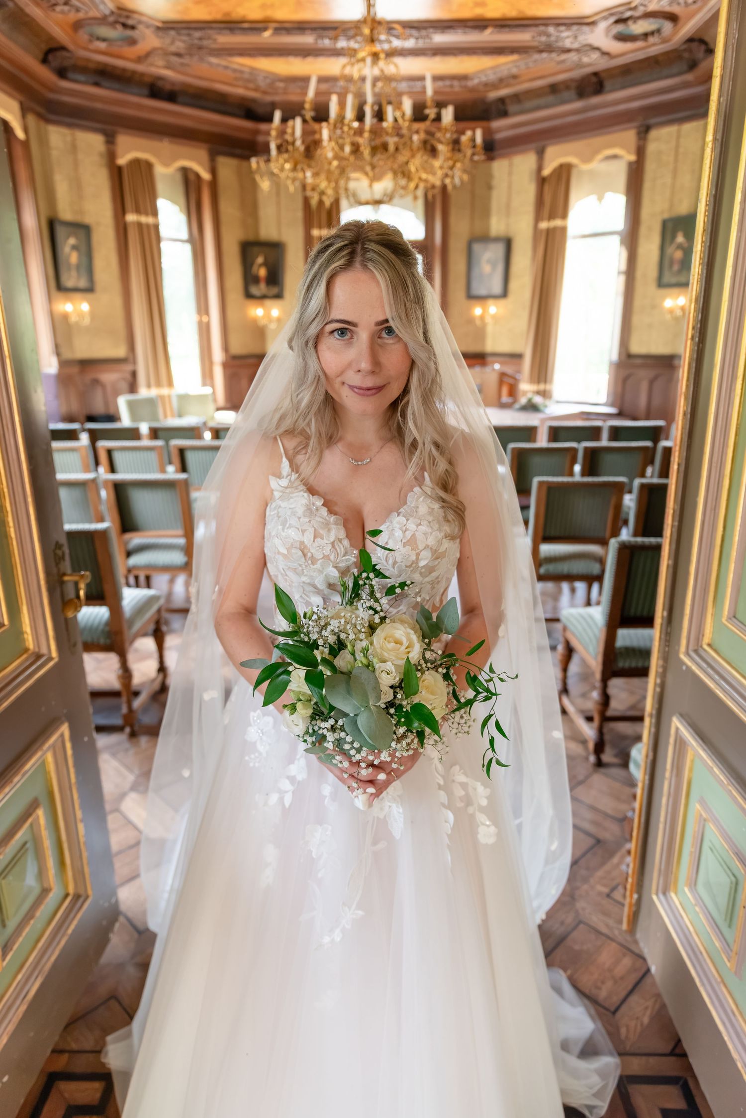 Intieme foto van een bruid in witte trouwjurk die haar boeket vasthoudt terwijl ze in de deuropening van de klassieke trouwzaal staat.